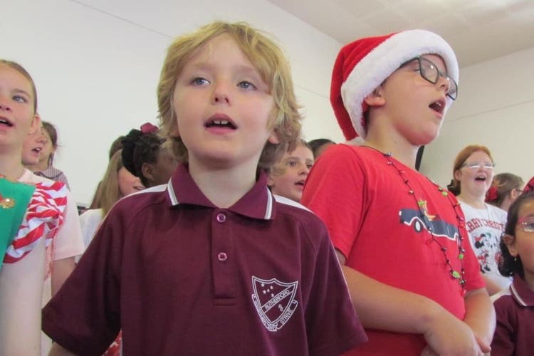 2 boys are in the foreground with their mouths open, singing. The boy on the left wears his maroon school shirt and the boy on the right wears a red shirt and Santa hat.
