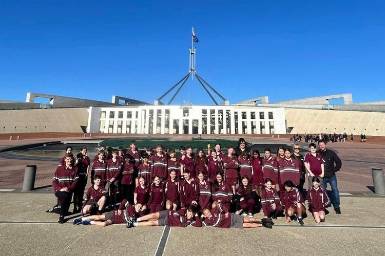 a large group of students and some teachers stand in front of Parlament House in Canberra. Three boys are laying on their sides, on the ground in front of the group with their hands propping their heads up.