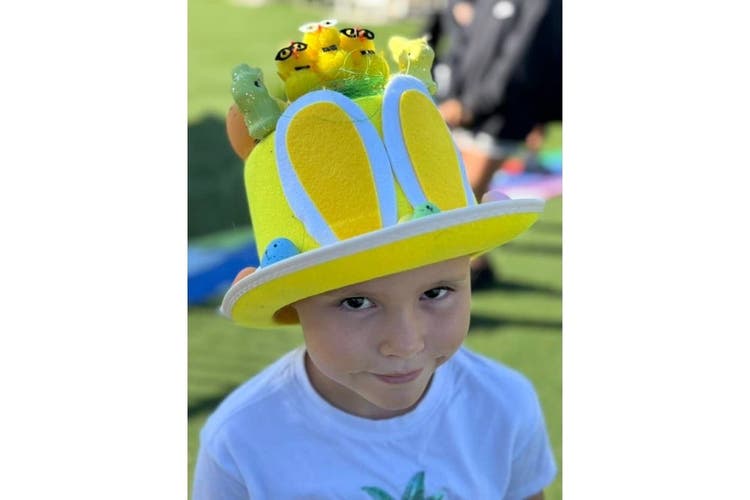 A child looks up at the camera with a small smile. They are wearing a bright yellow hat with bunny ears and yellow chicks on it.
