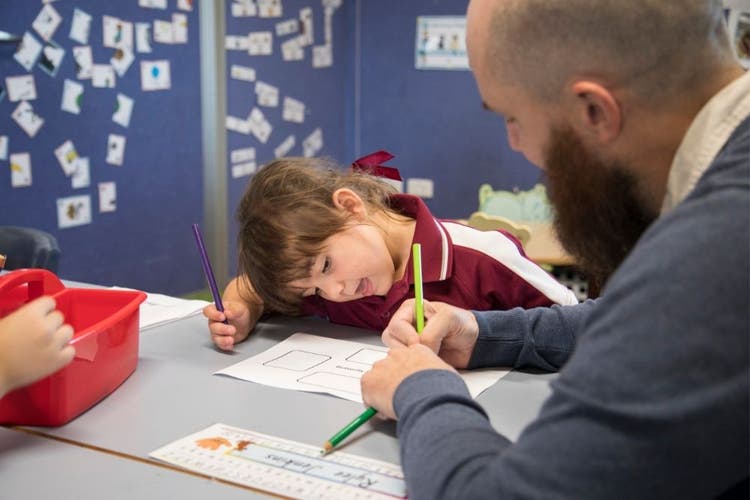 In the foreground, we see the side of a male teacher as he writes on some paper while a child leans forward to look at what he is doing.