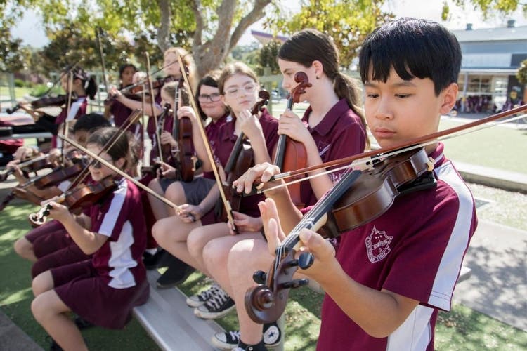 A group of students holding violins chat in the background while a boy plays the violin in the foreground.