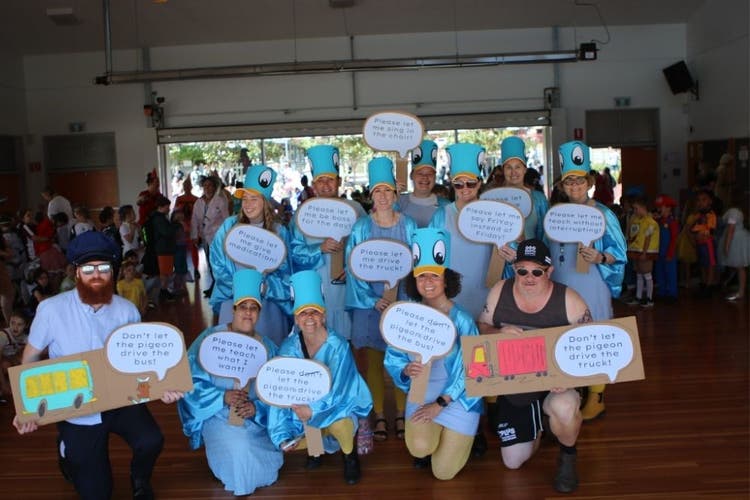 A group of teachers a dressed in costumes for Book Week. The females are wearing light blue and have cardboard pigeon heads for hats. There is a man to the right who is dressed as a bus driver and holds a cardboard bus and a man to the right is dressed in a singlet, shorts and cap and holds a cardboard truclk. Everyone holds speech bubble shaped cardboard signs with quotes such as,