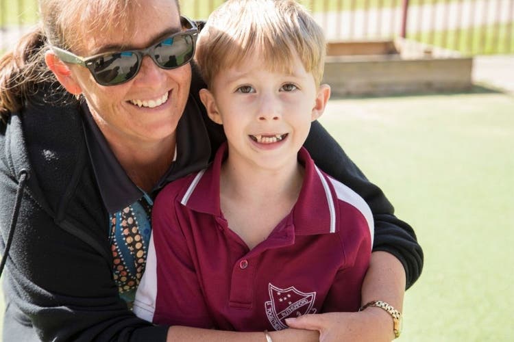 A teacher with sunglasses on has her arms around a child hugging him from behind as they both look directly at the camera with large smiles.