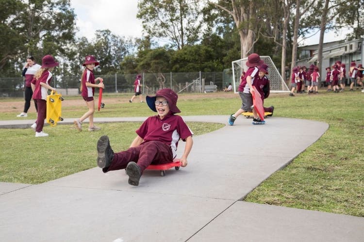 A child has a large smile on his face as he rides a red scooterboard along a path with his legs raised.