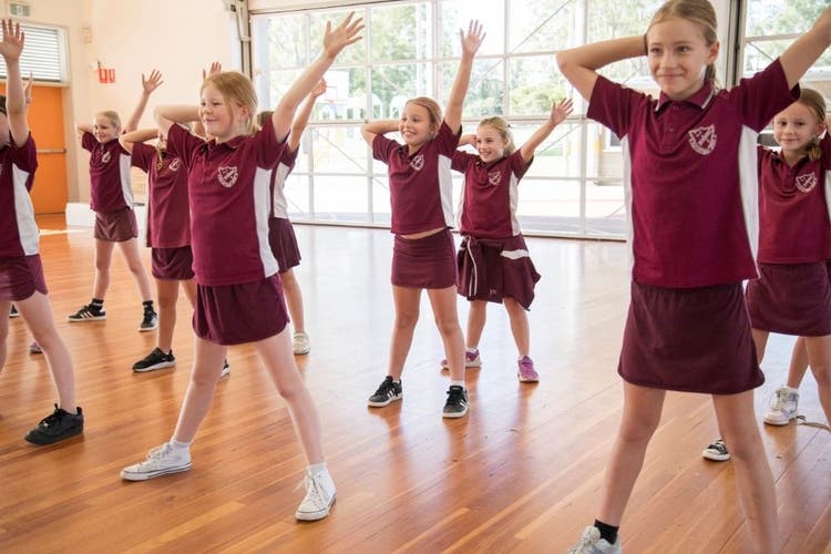 Students are dancing, standing in rows with one hand on behind their heads and the other hand raised.