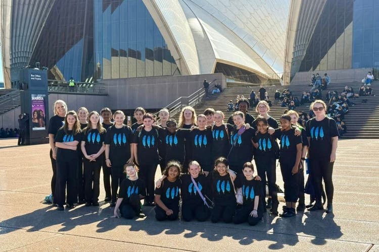 A group of students and 2 teachers stand in front of the Sydney Opera House. The sails of the builind can only be partially seen in the background. The students and teachers all wear black shirts with three blue feathers printed on them.