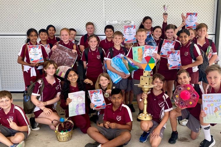 A large group of students hold props, certificates and sa trophy with a large golden dice on top all look at the camera and are smiling. A child at the back of the group holds a trophy in the air with one hand.