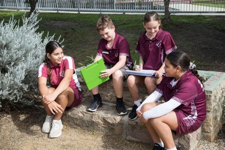 4 students sit on a sandstone block. The boy holds a green clipboard and looks at it, smiling, while the 3 girls talk to each other.