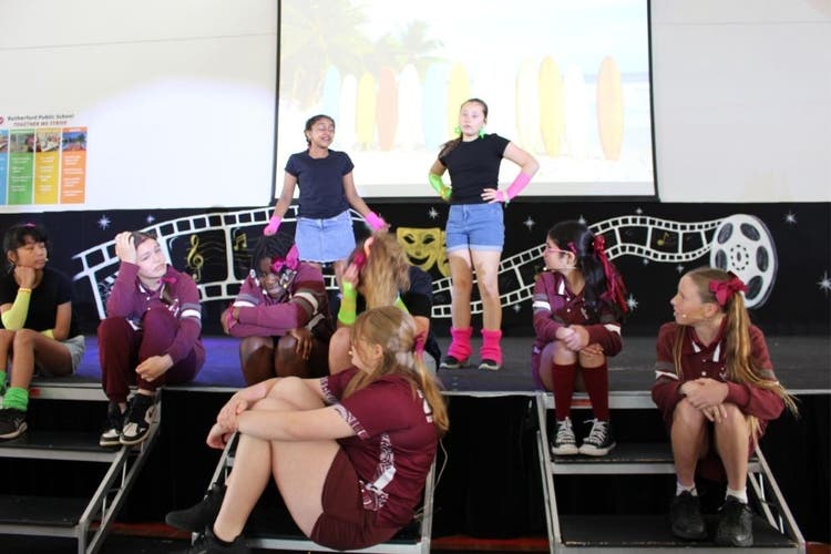2 girls stand on stage while a group of girls sit around them, looking up at them. One girl has her hands on her hips while the other holds her hands out to the side as she talks to the group.