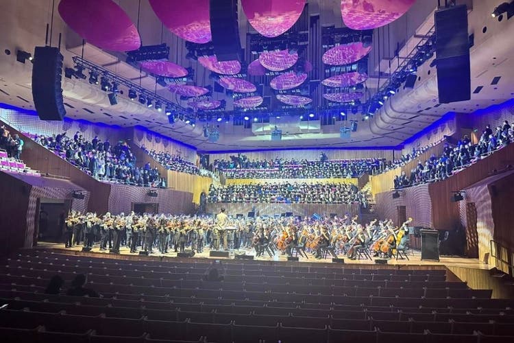 A photo looking towards the stage of the Opera House where string students with string instruments can be seen seated on the stage. There are students seated around the stage too.