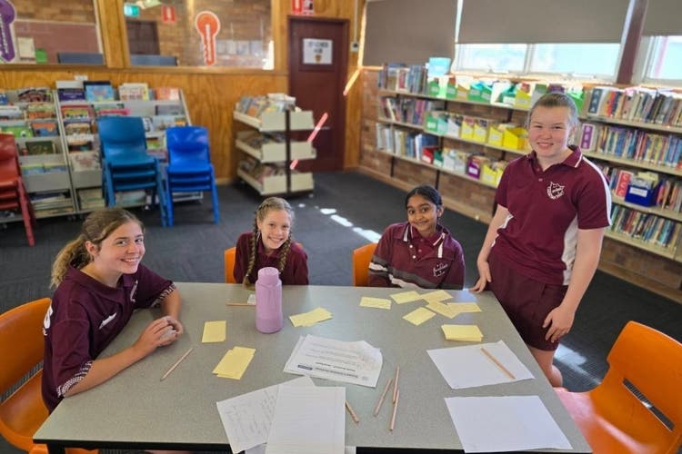 3 students sit at a table while a fourth student stands, They are all smiling and lookingn towards the camera. they have palm cards spread across the table and behind them there are books on book shelves as they are in a library.