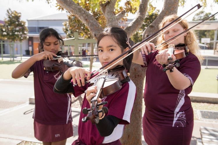 Three violin students stand under the shade of a tree playing their viiolins. The students on the left and right of the photo look at their violins as they play, while the child in the centre of the photo looks directly at the camera.