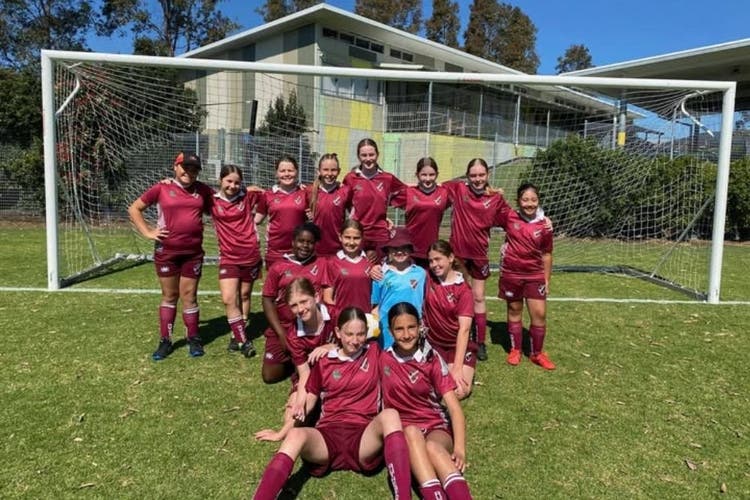 A group of girls soccer players are grouped together in front of a soccer goal. The back row are standing, the middle row are kneeling and 2 girls sit on the ground at the front.