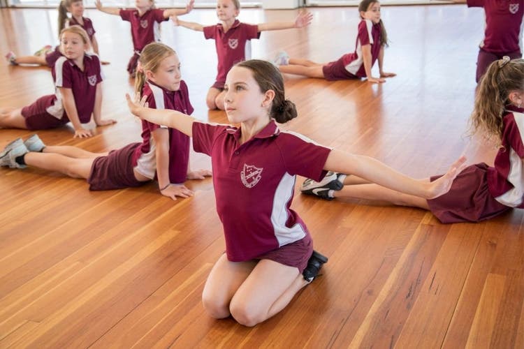 In the foreground, a girl is on her knees with her arms outstretched, looking into the distance. In the background, other dancers are holding poses. Some have their legs stretched out behind them while propping themselves up on their hands, while other students hold the same pose as the girl in the foreground.