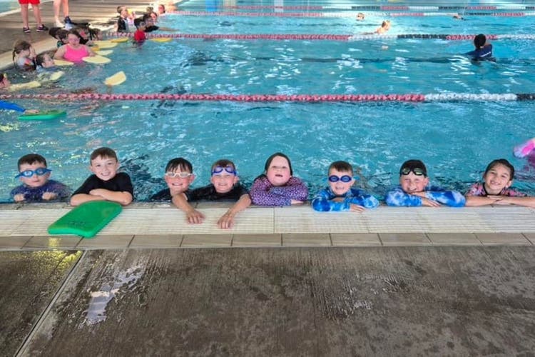 a group of eight students are in the water of a swimming pool and are resting at the pool edge, looking at the camer. One student has a green kickboard in under his arms.