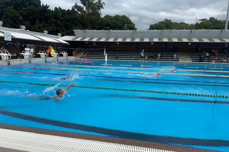 The photo looks across the pool and children can be seen swimming in the lanes. Water is splashing and arms can be seen.