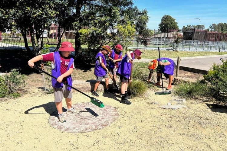 A group of 4 students sweep the ground in the Yarning circle as a teacher bends over to tend to a plant. teh students and the teacher are all wearing purple environment group vests.