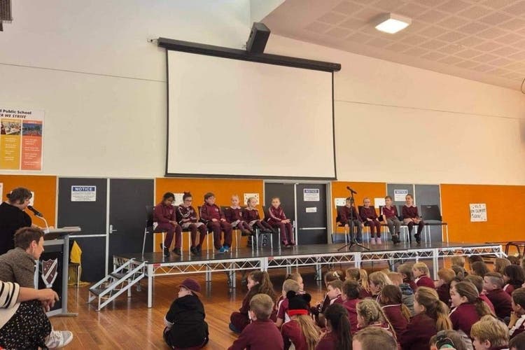 Students are seated in a row on a stage in the hall. A mircrophone is positioned in the centre of the stage. A teacher is to the left of the stage and is speaking to the students through a microphone. An audience can be seen seated in front of the stage.