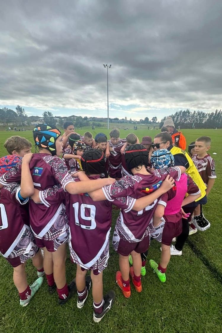 A group of rugby players stand in a circle with arms around each others shoulders. A teacher can be seen on the right talking to the group.