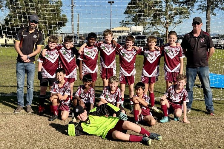 A boys soccer team pose in fron a soccer goal with their 2 coaches with the goal keeper laying on the ground with one hand propping up his head and the other on his hip.