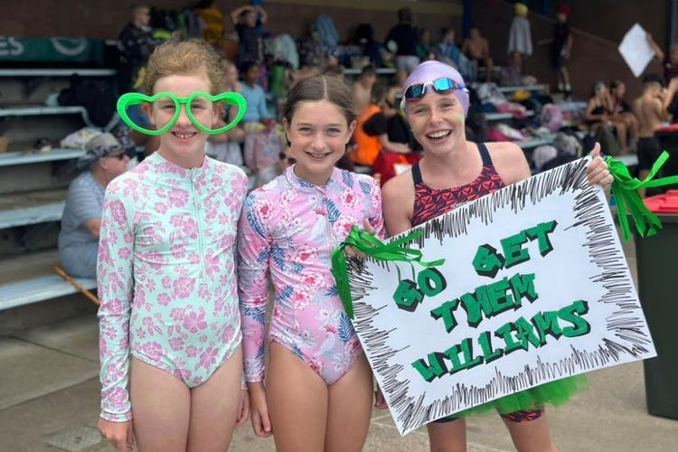 Three students dressed in swimwear, are smiling at the camera. The child on the left has large heart shaped green glasses on and the child on the right holds a sign that says,