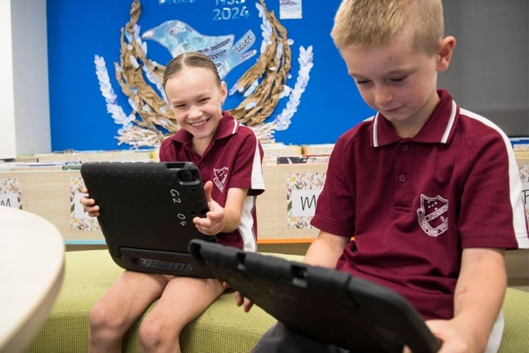 A boy looks at an iPad which he holds in his hands. Behind him a girl also holds an iPad, but is looking at the boy's iPad and is smiling at the screen.