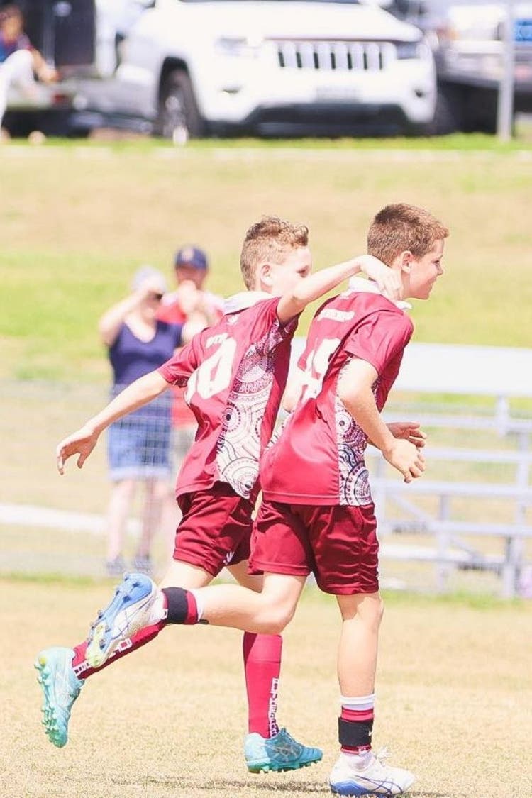 Two soccer player run across the field with their backs angled away from the camera. The boy on the left is reaching to put his arms around the shoulders of the boy on the right.