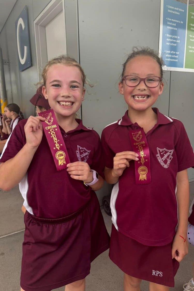 two students smile at the camera while holding maroon cross-country place ribbons.