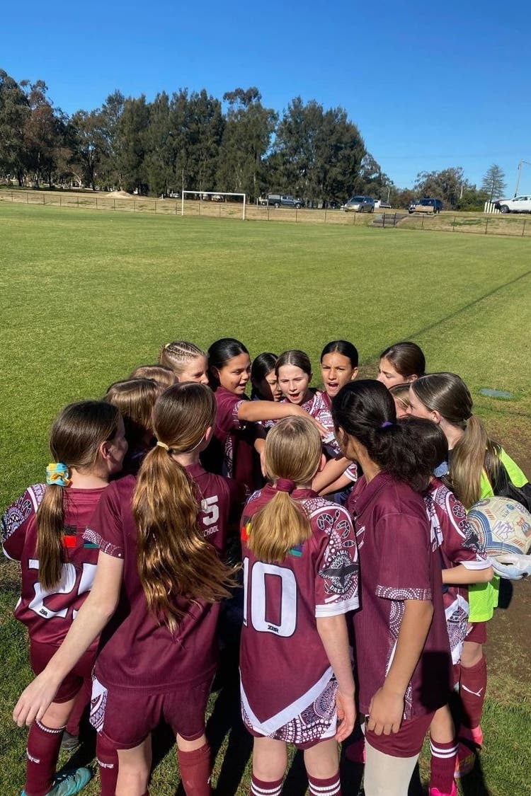A group of girl soccer player huddle together in a circle at the edge of a soccer field.