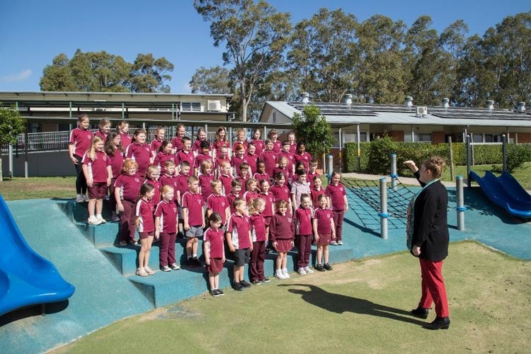The choir stand in rows on the steps of the blue amphitheatre which is based in sunshine. A teacher stands in front of the choir conducting them.